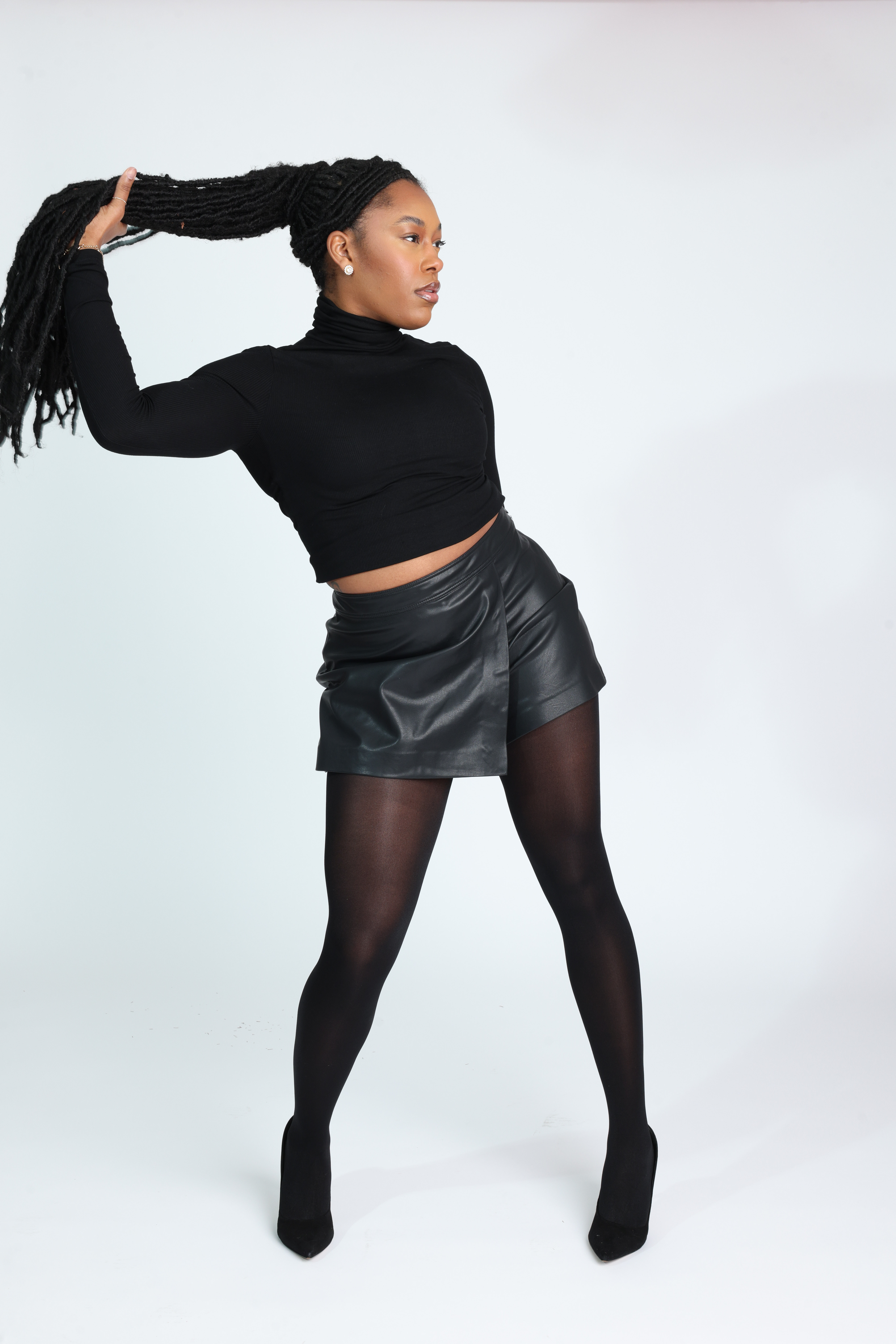 Confident Black woman in an all-black outfit — fitted turtleneck, leather mini skirt, sheer tights, and heels — sitting gracefully on a stool with hands in her hair. Minimalist studio shoot showcasing elegance, power, and poise. Editorial fashion inspiration for modern women embracing confidence and self-expression. Studio fashion portrait featuring a model in black minimalist styling seated on a stool against a white backdrop.