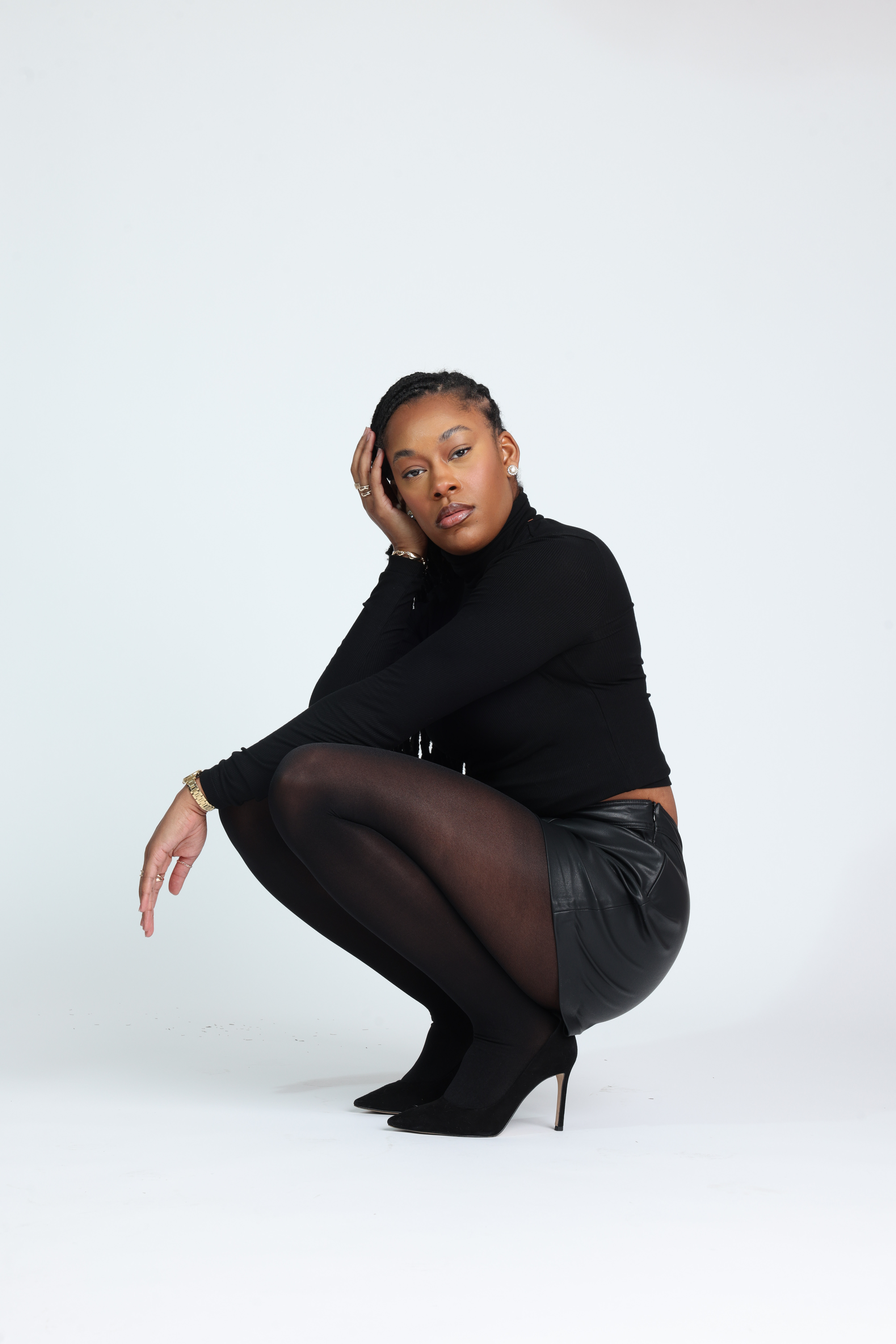 Confident Black woman in an all-black outfit — fitted turtleneck, leather mini skirt, sheer tights, and heels — sitting gracefully on a stool with hands in her hair. Minimalist studio shoot showcasing elegance, power, and poise. Editorial fashion inspiration for modern women embracing confidence and self-expression. Studio fashion portrait featuring a model in black minimalist styling seated on a stool against a white backdrop.