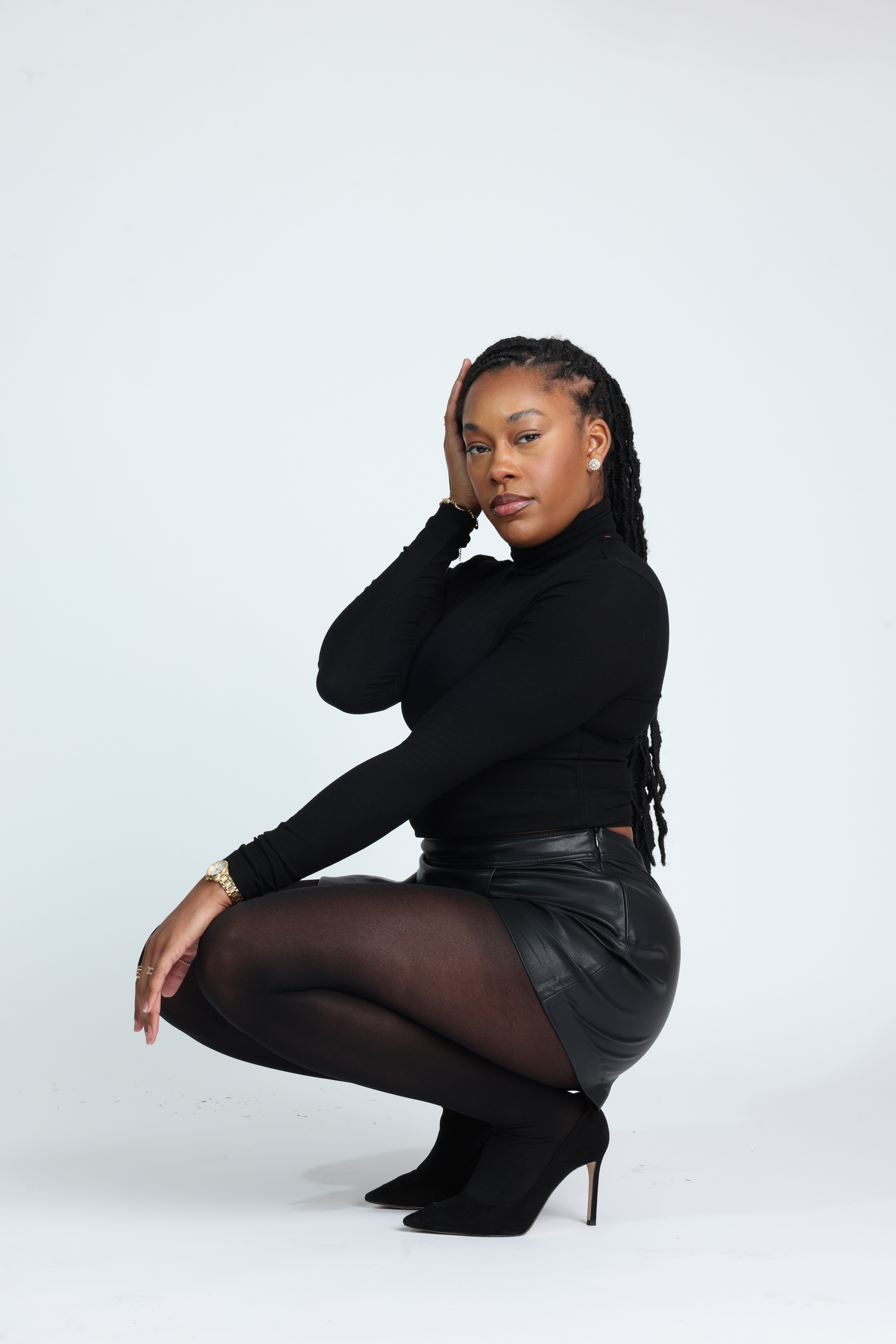 Confident Black woman in an all-black outfit — fitted turtleneck, leather mini skirt, sheer tights, and heels — sitting gracefully on a stool with hands in her hair. Minimalist studio shoot showcasing elegance, power, and poise. Editorial fashion inspiration for modern women embracing confidence and self-expression. Studio fashion portrait featuring a model in black minimalist styling seated on a stool against a white backdrop.