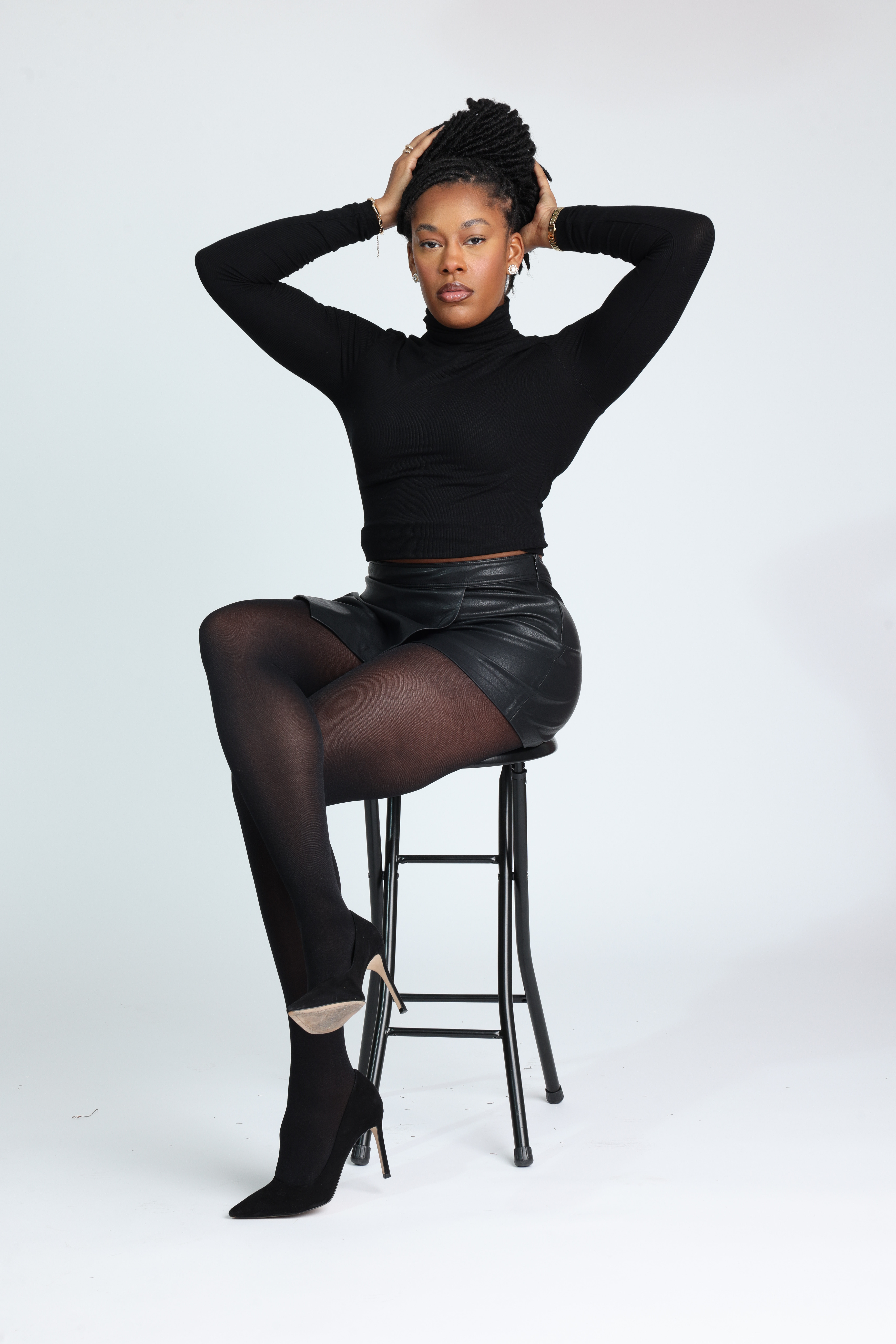 Confident Black woman in an all-black outfit — fitted turtleneck, leather mini skirt, sheer tights, and heels — sitting gracefully on a stool with hands in her hair. Minimalist studio shoot showcasing elegance, power, and poise. Editorial fashion inspiration for modern women embracing confidence and self-expression. Studio fashion portrait featuring a model in black minimalist styling seated on a stool against a white backdrop.